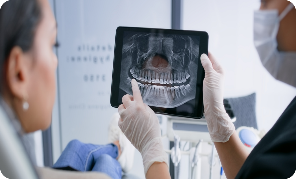 A dental professional is showing a patient a panoramic X-ray of their teeth on a tablet during a dental consultation.